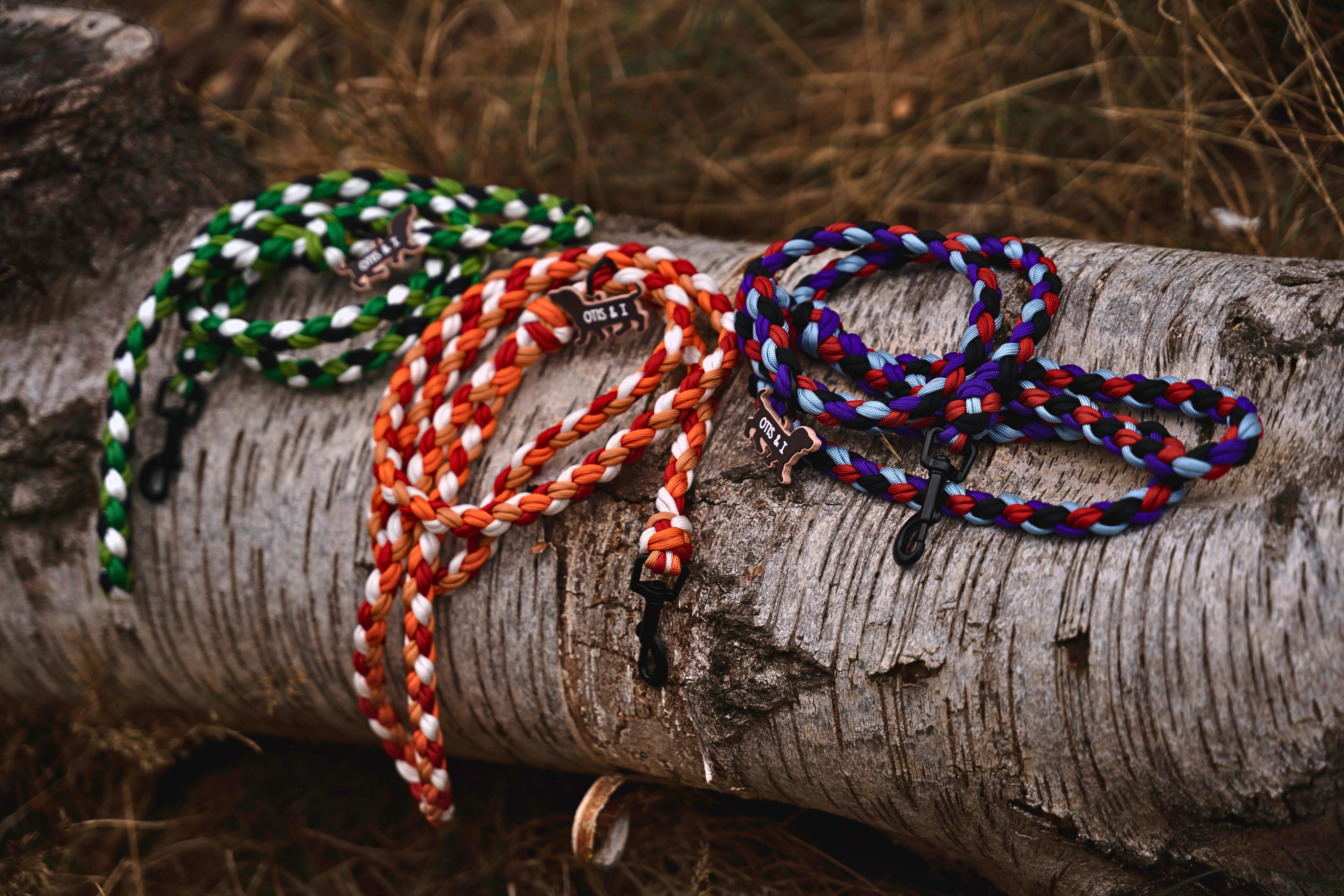 Three handcrafted braided dog leads in green, orange, and multicolor on a wooden log.